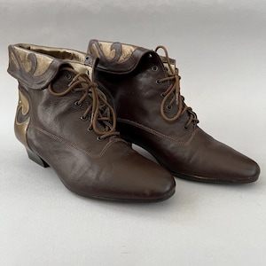 May include: A pair of dark brown leather lace-up ankle boots featuring a folded cuff detail with metallic gold accents. The boots have a rounded toe, a low heel, and matching brown laces threaded through metal eyelets along the front closure.