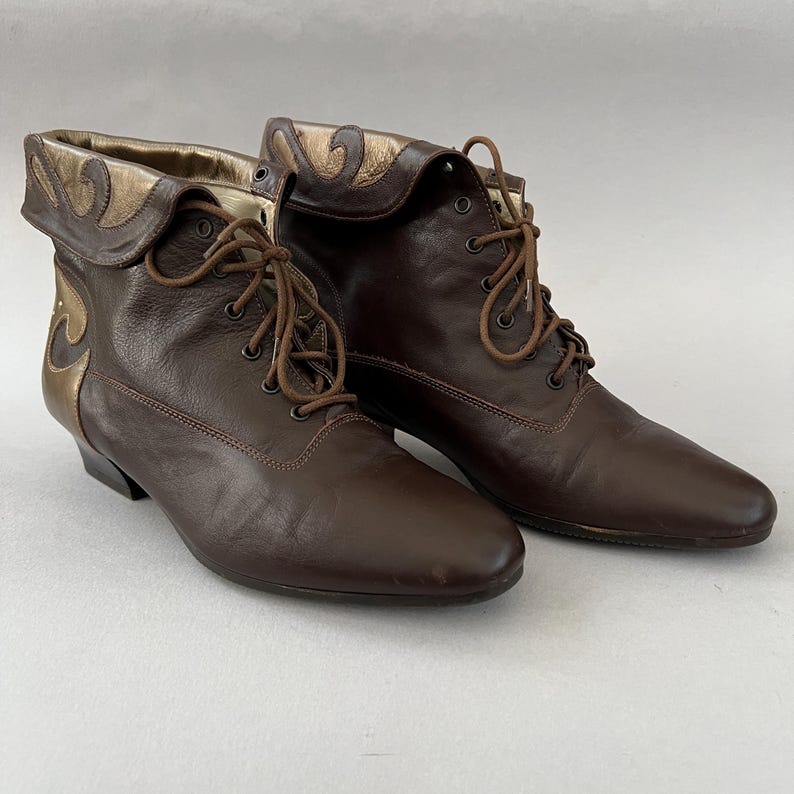 May include: A pair of dark brown leather lace-up ankle boots featuring a folded cuff detail with metallic gold accents. The boots have a rounded toe, a low heel, and matching brown laces threaded through metal eyelets along the front closure.