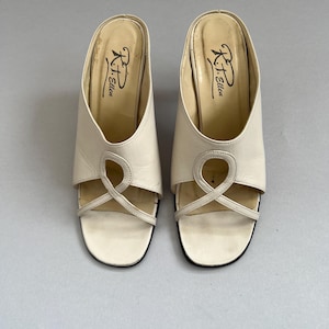 May include: A pair of off-white slip-on mules featuring a wide strap across the vamp with a decorative looped cutout design. The footbed is light beige and displays the black printed text R.T. Ellen near the heel area of each shoe.