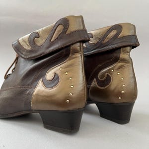 May include: A pair of ankle boots featuring a two-tone design in dark brown and metallic gold leather. The boots have a fold-over cuff, a low block heel, and decorative gold-tone studs arranged in a curved pattern along the side panels.
