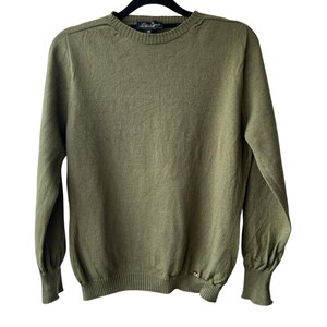 May include: An olive green long-sleeve sweater featuring a crew neckline, ribbed cuffs, and a ribbed hem. The garment includes a small gold-toned metal emblem near the bottom hem and a dark blue interior neck label with the text "La Ligne" and "M".