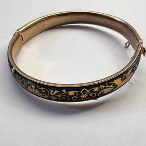 May include: A gold-coloured hinged bangle bracelet with a black enamel band. The black band features a gold floral design. The bracelet is closed and appears to be vintage.