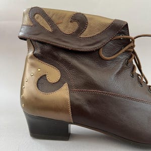 May include: A dark brown and metallic gold leather ankle boot featuring a fold-over cuff, decorative swirling leather overlays, brown laces with metal eyelets, and a low block heel. The side panel is accented with a vertical row of small gold studs.