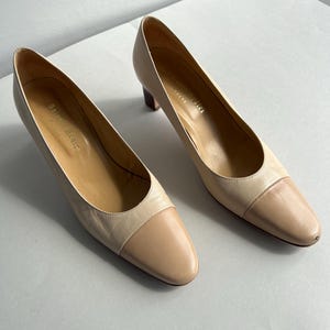 Könnte beinhalten: Ein Paar zweifarbige, beige Pumps aus Leder mit runder Spitze und kleinem Absatz. Die Zehenkappe hat einen helleren Beigeton als der Rest des Schuhs. Die Schuhe stehen auf einer weißen Oberfläche.