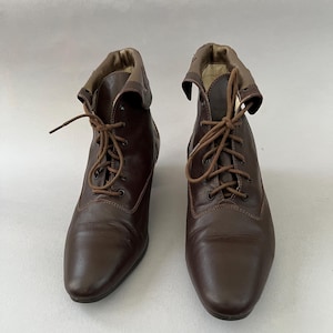 May include: A pair of dark brown ankle boots featuring a lace-up front with matching brown laces, a rounded toe, and a folded collar detail at the top. The boots have a smooth finish and a structured design with visible stitching along the vamp.