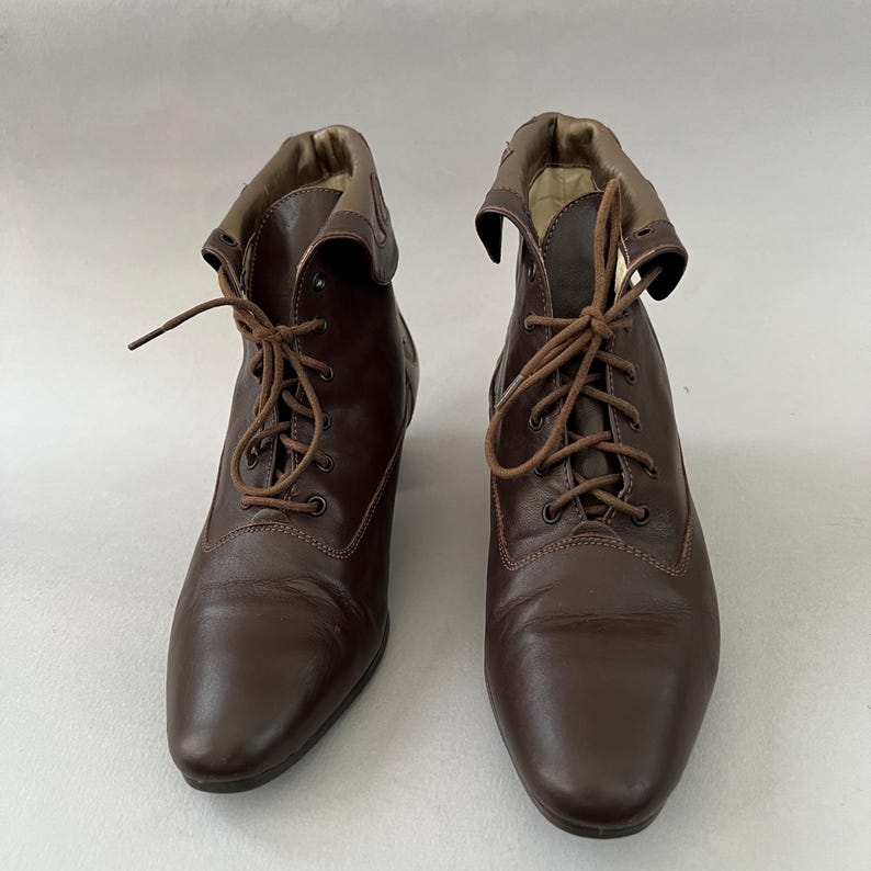 May include: A pair of dark brown ankle boots featuring a lace-up front with matching brown laces, a rounded toe, and a folded collar detail at the top. The boots have a smooth finish and a structured design with visible stitching along the vamp.