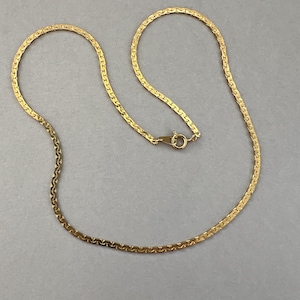 May include: A gold-colored necklace with a flat, herringbone chain design. The necklace has a small clasp and a delicate, elegant appearance. The chain is made of small, interlocking links, creating a smooth, flowing look. The necklace is displayed on a gray background.