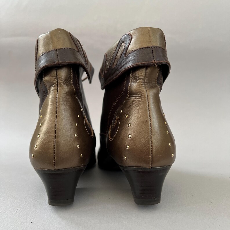 May include: A pair of ankle boots featuring a metallic bronze and dark brown leather construction. The boots have a folded cuff at the top, a low block heel, and are decorated with small gold-toned metal studs arranged in a curved pattern.