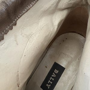 May include: The interior of a boot featuring a white leather lining and a white insole. A black rectangular label with the white text BALLY is attached to the heel area of the insole. The upper exterior edge of the boot is made of brown leather.
