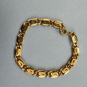 Può includere: Un braccialetto color oro con un design a maglie a forma di S ripetute. Il braccialetto è realizzato in un materiale metallico lucido e presenta una chiusura a fermaglio. Il gioiello è esposto su uno sfondo grigio.