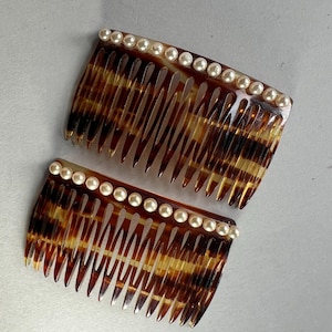 Könnte beinhalten: Zwei Vintage-Haarkämme mit Schildpattmuster und einer Reihe künstlicher Perlen am oberen Rand. Die Kämme haben feine Zähne und eine gebogene Form, geeignet zum Befestigen der Haare. Die Farbpalette umfasst Braun-, Bernstein- und Cremetöne.