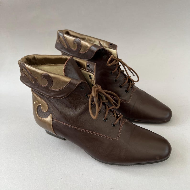 May include: A pair of dark brown ankle boots featuring a lace-up front with matching brown laces and eyelets. The boots have a folded cuff design with metallic gold accents along the collar and heel, showcasing a smooth finish and rounded toes.