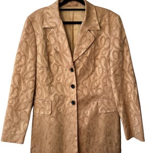 May include: A long-sleeved blazer in a metallic gold tone featuring a tonal paisley jacquard pattern throughout. The garment includes a notched lapel, three dark buttons down the center front, and two rectangular flap pockets on the lower front.
