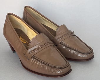 May include: A pair of taupe leather loafers with a low block heel and a braided strap detail across the vamp. The interior insole features the brand name BALLY printed in gold lettering. The shoes have a smooth finish and a rounded toe design.