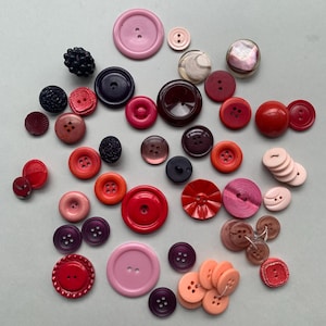 May include: A selection of vintage buttons in various sizes and shades of red, pink, purple, and brown. The buttons have different textures and designs, including smooth, ridged, and textured surfaces. Some buttons have two or four holes, while others have a shank.