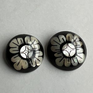 Puede incluir: Un par de pendientes negros y blancos con un diseño floral. Los pendientes están hechos de nácar y tienen un acabado brillante.