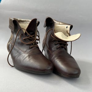 May include: A pair of dark brown leather ankle boots featuring a lace-up front closure with thin brown laces, a folded collar design, and a smooth, cream-coloured interior lining. The boots have a rounded toe and a low, integrated heel structure.