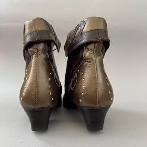 May include: A pair of ankle boots featuring a metallic bronze and dark brown leather construction. The boots have a folded cuff design, small gold-tone metal studs arranged in a curved pattern on the heel, and a low, dark brown block heel.
