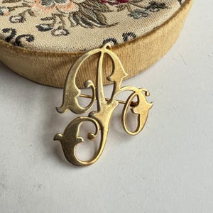 May include: A gold-toned vintage brooch in the shape of the letters "AF" in a decorative script. The brooch has a pin closure on the back. The brooch is set against a background of a floral embroidered fabric.