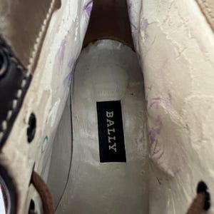 May include: The interior of a shoe featuring a white insole with a black rectangular label printed with the word BALLY in white capital letters. The inner side walls are white with faint purple markings and dark brown leather trim near the eyelets.