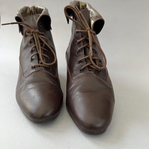 May include: A pair of dark brown leather ankle boots featuring a lace-up front with matching brown laces and metal eyelets. The boots have a folded-over collar design that reveals a metallic gold-toned interior lining and a rounded toe shape.