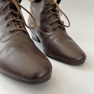 May include: A pair of dark brown leather lace-up boots featuring a rounded toe, a low heel, and matching brown laces threaded through metal eyelets. The boots have a smooth finish and a structured ankle height, showcasing a classic design.