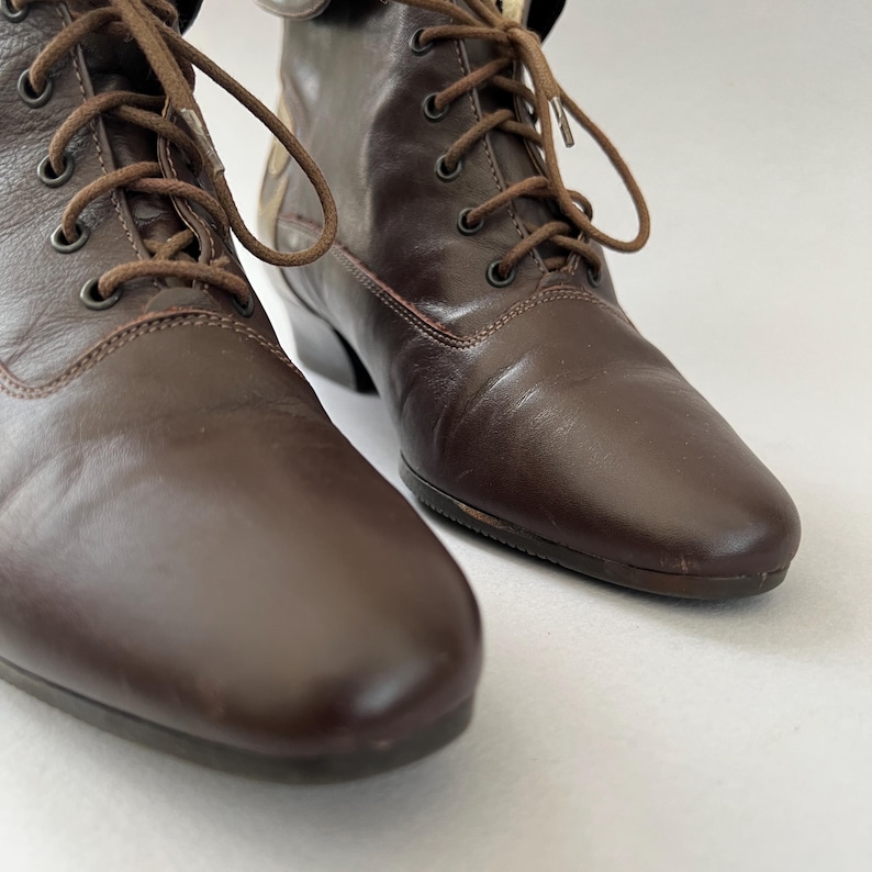 May include: A pair of dark brown leather lace-up boots featuring a rounded toe, a low heel, and matching brown laces threaded through metal eyelets. The boots have a smooth finish and a structured ankle height, showcasing a classic design.