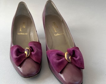 May include: A pair of plum-coloured leather pumps featuring a suede bow on the toe with a gold-tone accent. The interior of the shoes is lined in a light tan material and features the text Bally Switzerland bellezza printed in gold lettering.