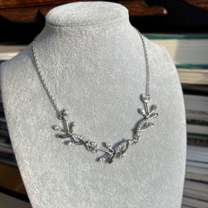 May include: Silver-toned necklace with a leaf design. The necklace features delicate, stylized leaves adorned with small, clear crystals. The necklace is displayed on a gray mannequin head, showcasing its elegant design.