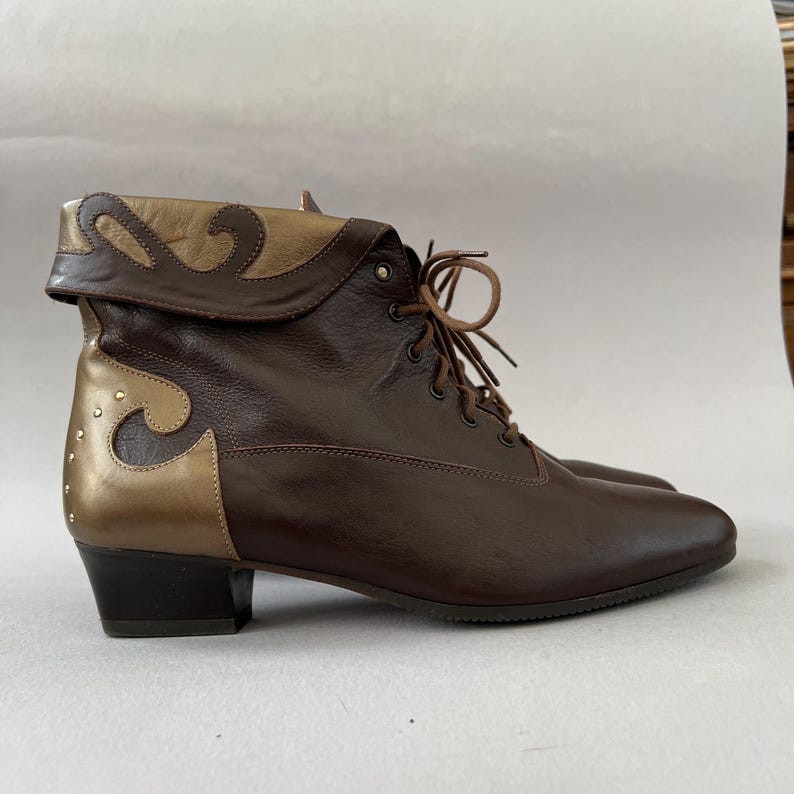 May include: A dark brown leather ankle boot with a lace-up front and a low block heel. The boot features a fold-over cuff and heel panel in a metallic gold finish with decorative swirling cutouts and small silver-toned studs along the back heel.