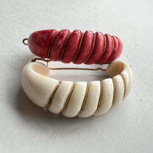 Op de afbeelding: Twee decoratieve haarspelden. Eén is felrood, de andere is gebroken wit. Beide zijn spiraalvormig en omwikkeld met goudkleurig draad. De haarspelden staan tegen een lichte achtergrond.