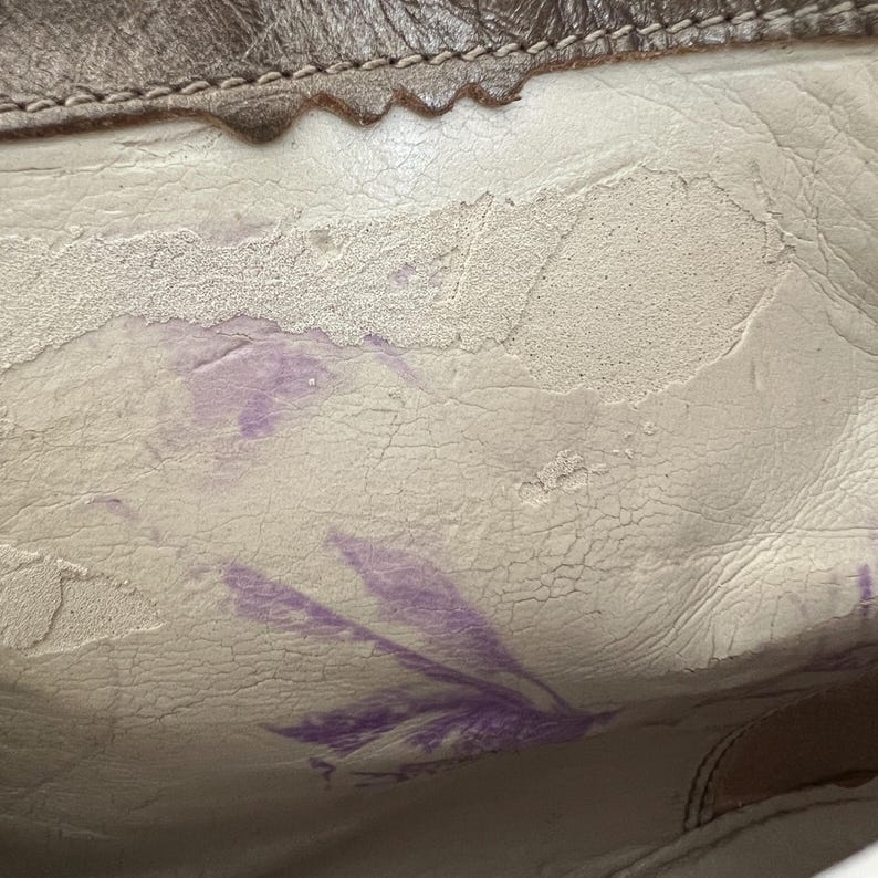 May include: A close-up view of the interior of a bag featuring a cream-coloured lining with a visible cracked texture and signs of wear. The material shows areas of peeling and light purple ink-like stains across the surface, with a brown leather trim.