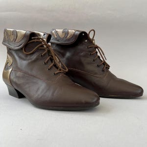 May include: A pair of dark brown leather ankle boots featuring a lace-up front closure with matching brown laces. The boots have a fold-over collar design with metallic gold leather accents along the top edge and the back heel area.