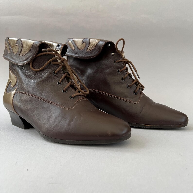 May include: A pair of dark brown leather ankle boots featuring a lace-up front closure with matching brown laces. The boots have a fold-over collar design with metallic gold leather accents along the top edge and the back heel area.
