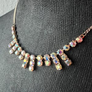 Puede incluir: Un collar plateado con brillantes iridiscentes, redondos y rectangulares. El collar tiene una cadena delicada y un diseño tipo babero, con los brillantes dispuestos en un patrón en cascada. El collar se muestra sobre una superficie negra.