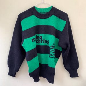 Pode incluir: Camisola de malha listrada em preto e verde com o texto "ming mchting" em preto.