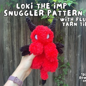 Puede incluir: Un juguete de peluche de imp crochetado rojo y negro con puntas de lana esponjosas. El juguete está siendo sostenido por una mano. El texto "LOKI THE IMP SNUGGLER PATTERN WITH FLUFFY YARN TIPS! *DIGITAL DOWNLOAD ONLY!*" es visible en la imagen.