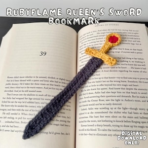 Op de afbeelding: Een handgemaakt gehaakt bladwijzer in de vorm van een zwaard, met een grijze handgreep en een gele en rode pareerstang. De bladwijzer ligt in een open boek, met de tekst "RUBYFLAME QUEEN'S SWORD BOOKMARK" zichtbaar.