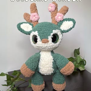 Può includere: Un giocattolo di cervo di peluche fatto a mano nei toni del verde, bianco e marrone, con grandi occhi espressivi. Il cervo ha corna decorate con fiori rosa e il testo "Willow The Woodland Wonder" sopra. Il testo "Pattern by @crochetwithkelsea on Instagram CrochetWithKelsea on Etsy" è sotto.