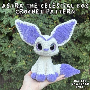Könnte beinhalten: Ein gehäkeltes Fuchskuscheltier aus weißem und violettem Garn mit großen Ohren, dunklen Augen und einem buschigen Schwanz. Der Text lautet ASTRA THE CELESTIAL FOX CROCHET PATTERN und DIGITAL DOWNLOAD ONLY. Der Fuchs hat eine weiche, strukturierte Optik.