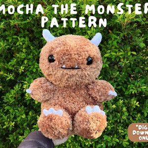 May include: A brown plush monster toy with white horns and a toothy grin. The toy is sitting on a hand in front of a green leafy background. The text "MOCHA THE MONSTER PATTERN" is at the top of the image and the text "*DIGITAL DOWNLOAD ONLY*" is at the bottom of the image.