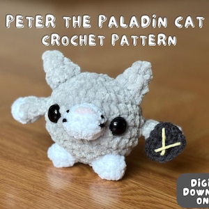 May include: A gray crocheted cat with white paws and a black crocheted shield. The cat has black eyes and a white nose. The text "PETER THE PALADIN CAT CROCHET PATTERN" is above the cat. The text "DIGITAL DOWNLOAD ONLY!" is below the cat.