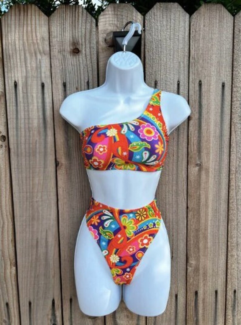 May include: One-shoulder swimsuit with a high-cut leg. The swimsuit is red with a colorful paisley pattern.