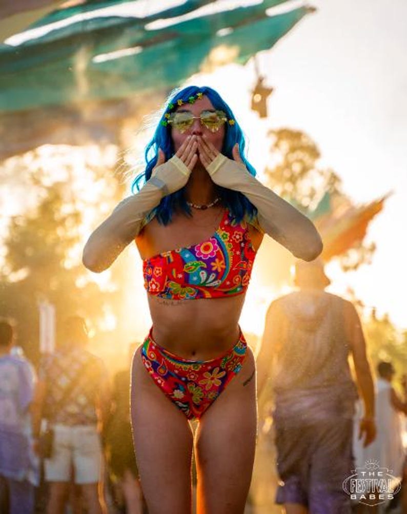 May include: A woman with blue hair and sunglasses is wearing a colorful bikini top and matching bottoms. She is covering her mouth with her hands and smiling. The background is blurry and shows a crowd of people.
