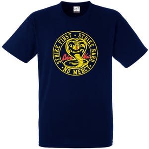 May include: Navy blue t-shirt with a yellow and red Cobra Kai emblem. The emblem features a cobra, the words "Strike First, Strike Hard, No Mercy," and the Cobra Kai name. The shirt is a short-sleeved crew neck.