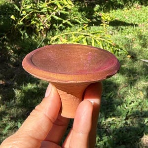 May include: A handmade, terracotta-colored ceramic bird bath with a wide, shallow bowl and a short, pedestal base. The bowl has a reddish-brown hue with a pinkish rim. The bird bath is held by a hand against a backdrop of green foliage.