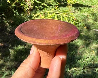 May include: A handmade, terracotta-colored ceramic bird bath with a wide, shallow bowl and a short, pedestal base. The bowl has a reddish-brown hue with a pinkish rim. The bird bath is held by a hand against a backdrop of green foliage.