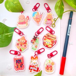 May include: A selection of vibrant acrylic keyrings with red clasps. The keyrings showcase diverse cartoon characters and food-themed designs, including a monkey with strawberries, a bunny holding a sign that says "Skittish", and a character in a jar.