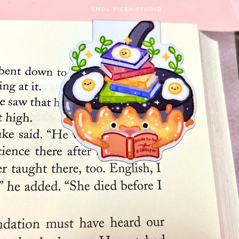 May include: A colorful bookmark featuring a cartoon crab holding a book titled "guide to be a calcifer". The crab is sitting in a frying pan with a stack of books and three fried eggs.