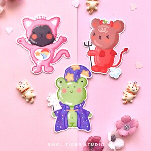 May include: Three colorful stickers featuring a pink cat wearing a rainbow costume, a red devil mouse, and a green frog wearing a purple wizard robe. The stickers are on a pink background.